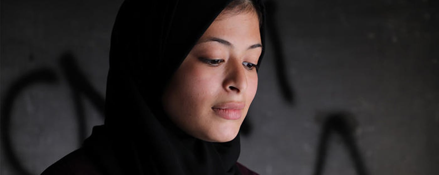 Portrait of Omaia, a 16-year-old Palestinian girl. She is standing in front a grey wall, looking down.