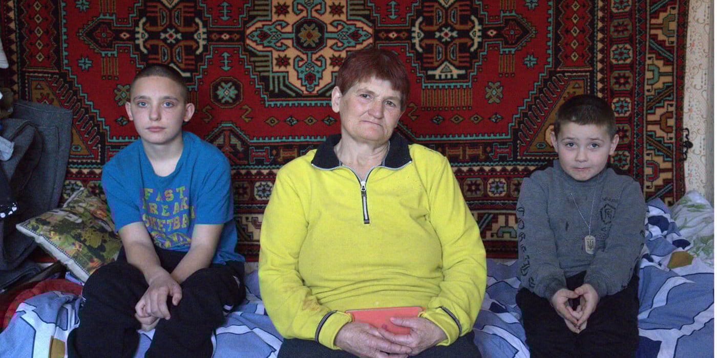 En äldre kvinna sitter mellan två pojkar på en säng i ett enkelt rum.