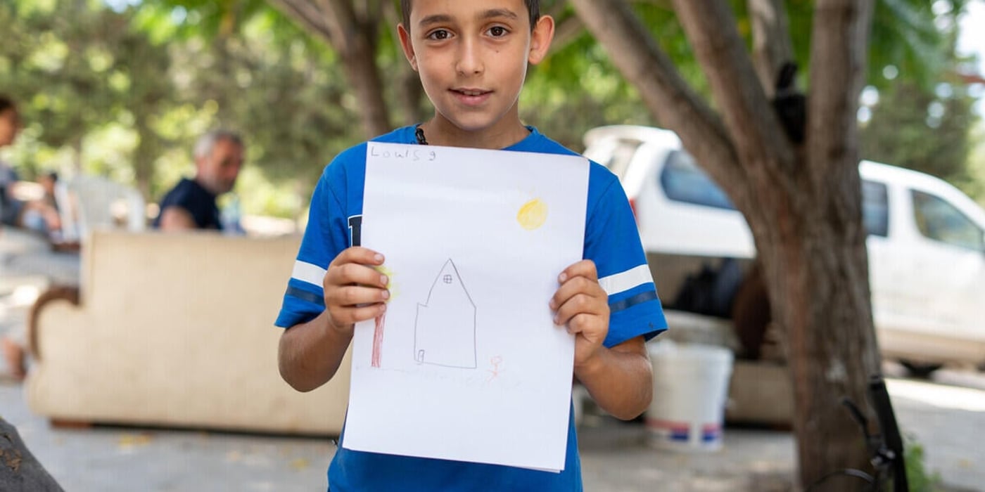 En pojke står utomhus och håller upp en teckning av ett hus med sol och en person. I bakgrunden syns träd, några vuxna och parkerade fordon.