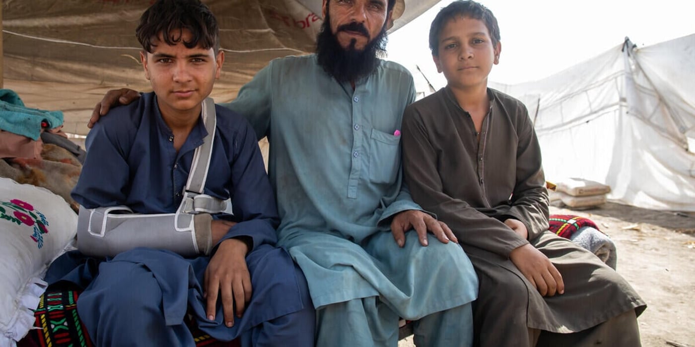 En man sitter i ett tält tillsammans med två pojkar. En av pojkarna har en armskena.  De befinner sig i ett tillfälligt boende efter jordbävningen.