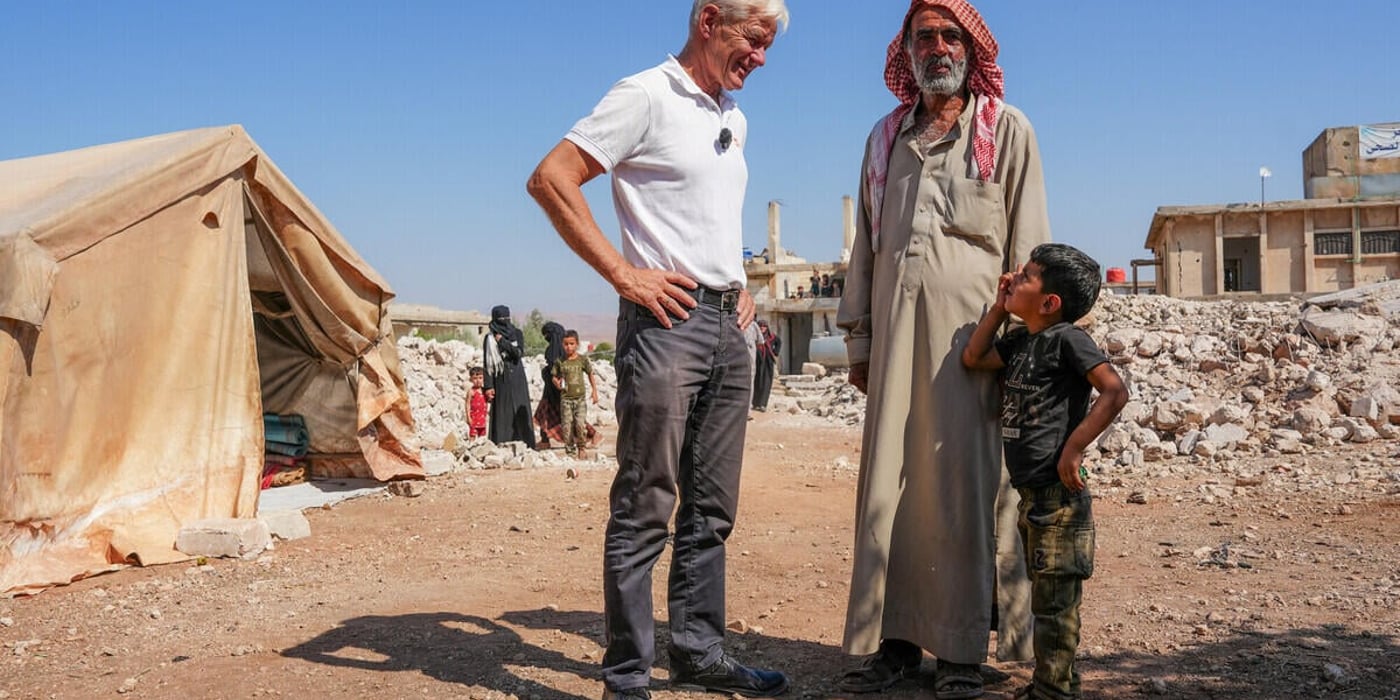 En man från Norwegian Refugee Council står och samtalar med en äldre man och ett barn i ett område med tält och förstörda byggnader. Bakom dem syns fler människor och rester av hus i det torra landskapet under en klar himmel.