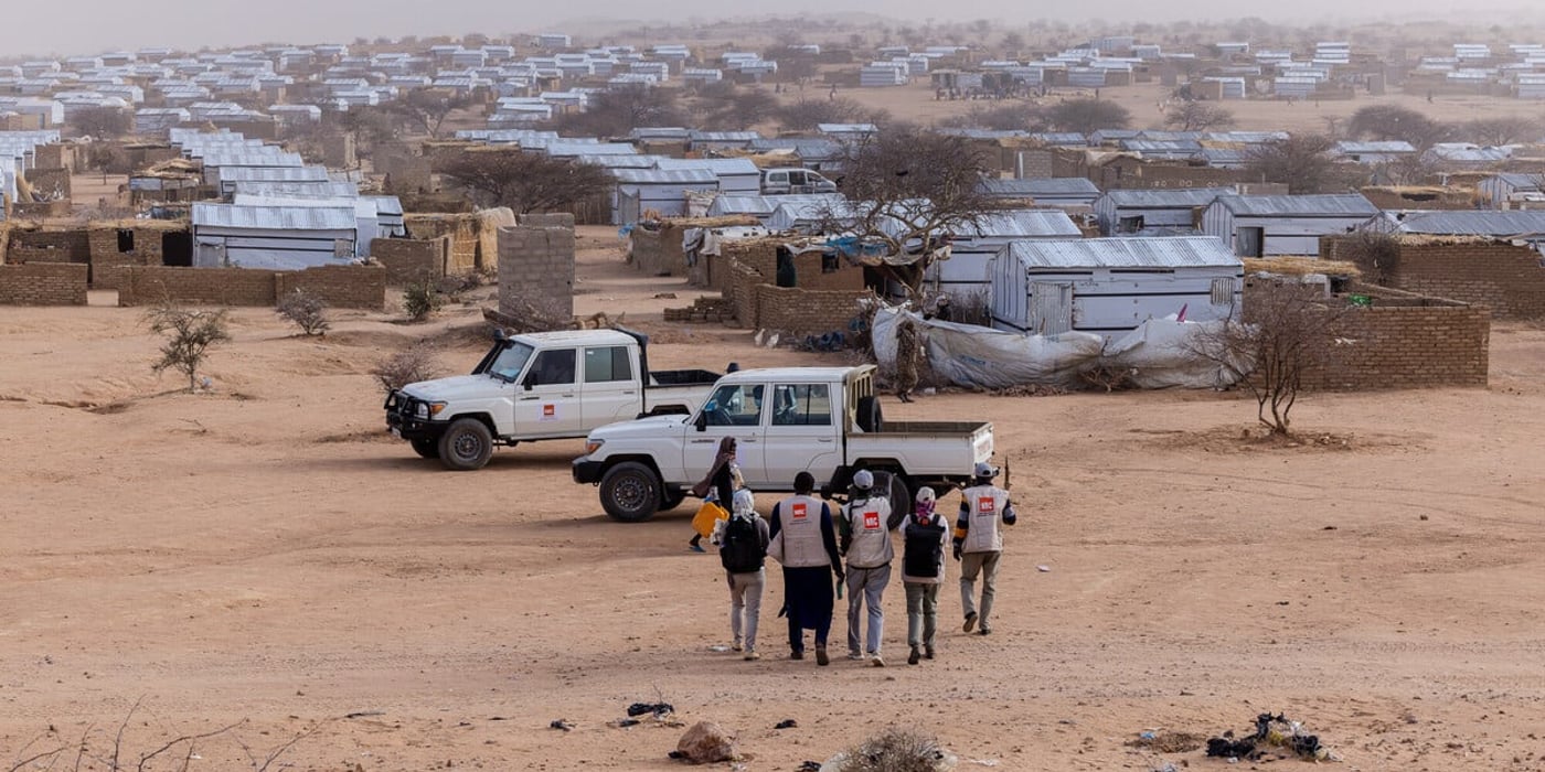 Hundratals enkla bostäder med plåttak breder ut sig över området och omges av sandig mark med gles vegetation. I förgrunden går en grupp humanitära medarbetare mot två terrängfordon parkerade i lägret.