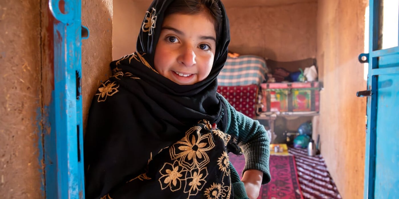 En ung flicka står i dörröppningen till sitt hem och ler mot kameran. Hon bär en svart sjal med broderade blommor i guld och en grön tröja.