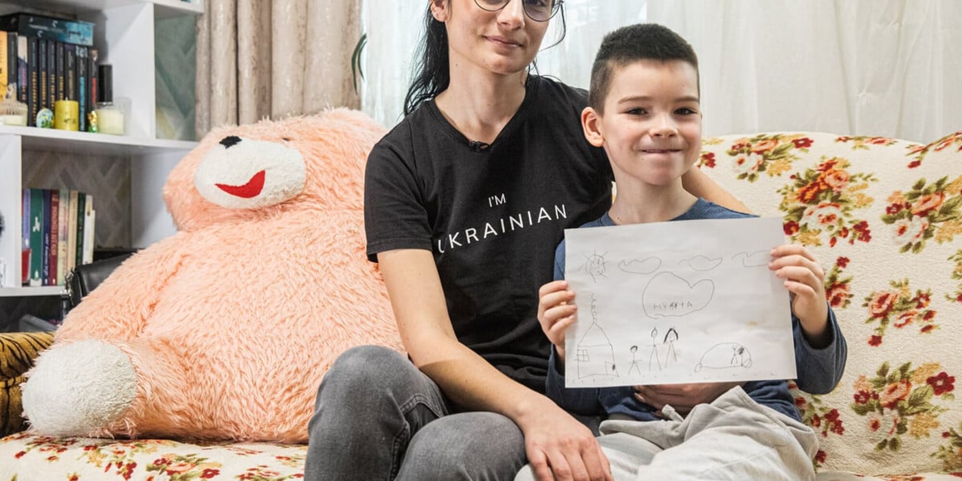 En kvinna och en pojke sitter tätt intill varandra i en soffa i ett vardagsrum. Kvinnan har armen om pojken och ser lugnt in i kameran.