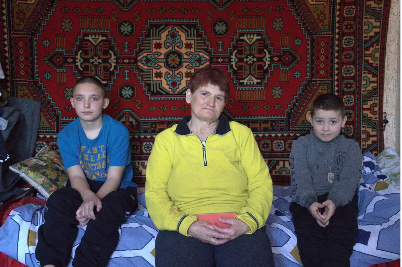 En äldre kvinna sitter mellan två pojkar på en säng i ett enkelt rum.