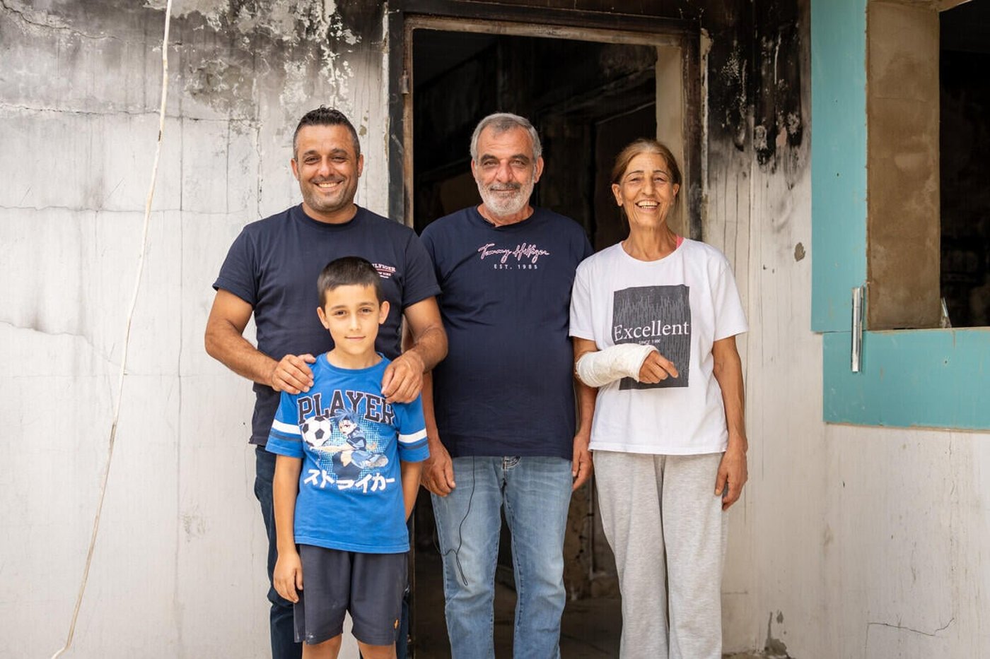 Fyra personer står tätt tillsammans framför en brandskadad byggnad. En vuxen håller en hand på pojkens axel, och alla ser mot kameran.
