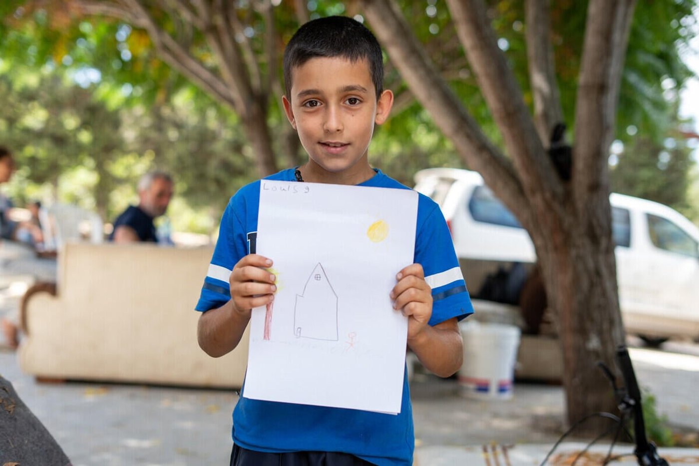 En pojke står utomhus och håller upp en teckning av ett hus med sol och en person. I bakgrunden syns träd, några vuxna och parkerade fordon.