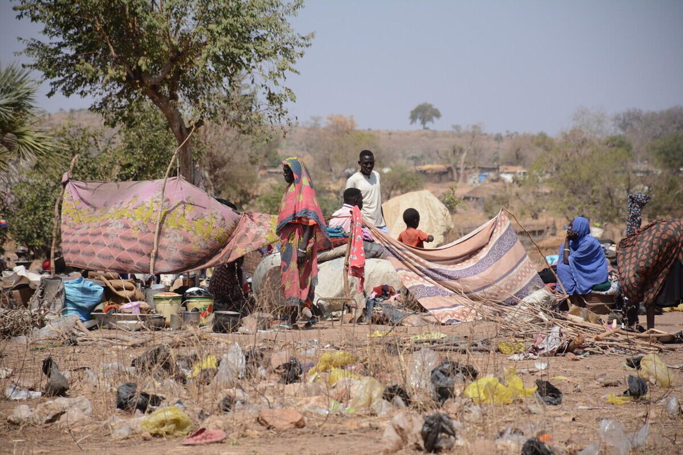En grupp människor står och rör sig runt provisoriska skydd byggda av filtar, tyg och grenar i ett torrt landskap.