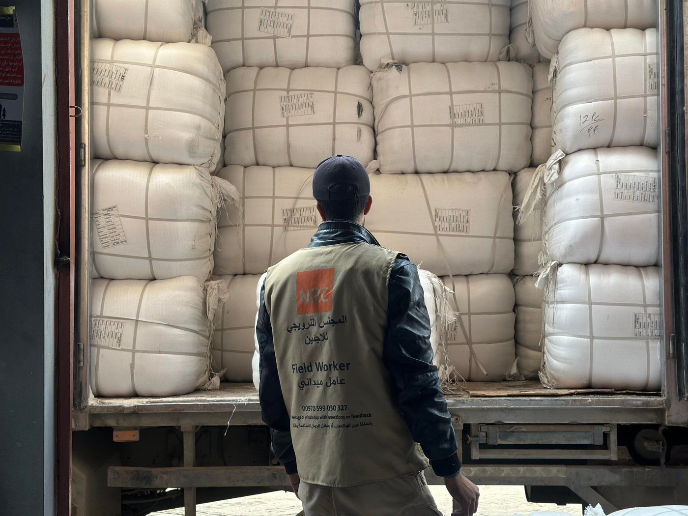 En man står bakom en lastbil lastad med stora vita säckar, staplade tätt intill varandra. Han bär en väst märkt med Norwegian Refugee Council och texten – Field Worker – och ser ut att inspektera eller förbereda lossning av humanitärt bistånd.
