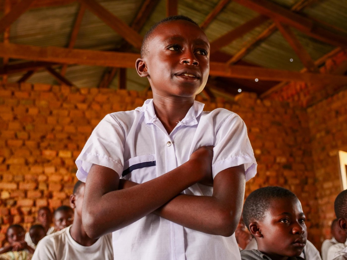 A boy standing up in a classroom. His arms are crossed and he is smiling.
