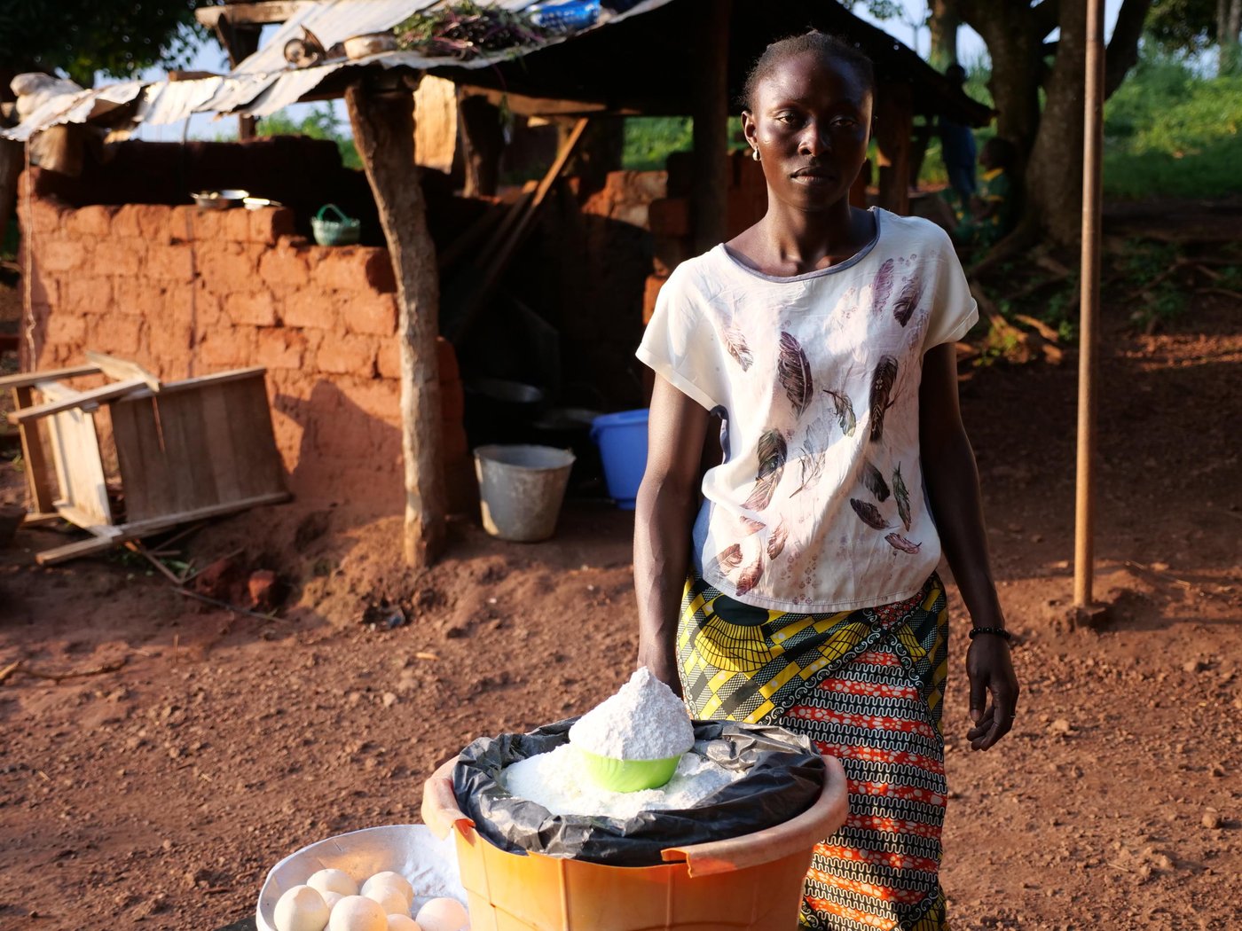 Nuela stands outside of her home in Boali, Central African Republic and shows off the items that she has for sale.
Nuela Ngaiboma is a 29 year old woman originally from Birlo village in Central African Republic. During CAR’s post electoral violence in December 2020, armed groups attacked Nuela’s village, Birlo. She fled and after an arduous journey she made it to Boali to stay with her aunt. In Boali, she received cash assistance from NRC to get back on her feet. She lives with her three childre, (two sons and a daughter) and sells food outside her house in Boali.
“When I was in the fields, I saw many people had started to run. When I asked why they were running, they said an armed group had entered the village and they had taken control of the village. I asked those running if they had seen my children. They said they had just fled as quick as they could. I had no choice but to go back and try to find my children. They tried to stop me from going to the village saying it was too dangerous. I told them I can’t abandon my children, and that I’m going to look for them so we can all stay together in the bush. When I returned to the village, I saw the armed men had taken the village. I heard gunshots coming from all directions. There were no villagers anywhere, there were not even any animals. I found my children and I ran with them to the bush until I reached the edge of a stream. When my aunt here in Boali learned about the attack, She called me and told me to come to Boali with a taxi-moto. I told her that there were no taxi-motos anywhere. How am I going to make it all the way to Boali? So my aunt asked some young people from Boali, to come to try to find me. They crossed through the bush on foot and they found me. It was around 9am when they found me and so we crossed through the bush together. It was around 4pm when we arrived here in Boali. As soon as my aunt saw me, she started to cry. She asked me to stay here and that’s why I’m here to this day. While I was here, NRC came here. They provided me with around $90 in cash in assistance. This helped to reduce my hardship here a little. One of my challenges right now is that my children are not going to school. They’re staying at home because their school is too dangerous. The children can’t go there because the school is surrounded by mines. I have many problems and I’m living in fear. I want there to be peace so my children can continue to go to school. I have so many worries on my mind. Because I am afraid. I am worried that what I just experienced might happen all over again. I just want peace for my country. So that we can work in our fields, go about our business, and see our children go to school while we live in peace. Then we will move past the memories of conflict and live in peace.” Nuela Ngaiboma, 29.
Photo: Itunu Kuku/NRC
Date: 13 July 2021