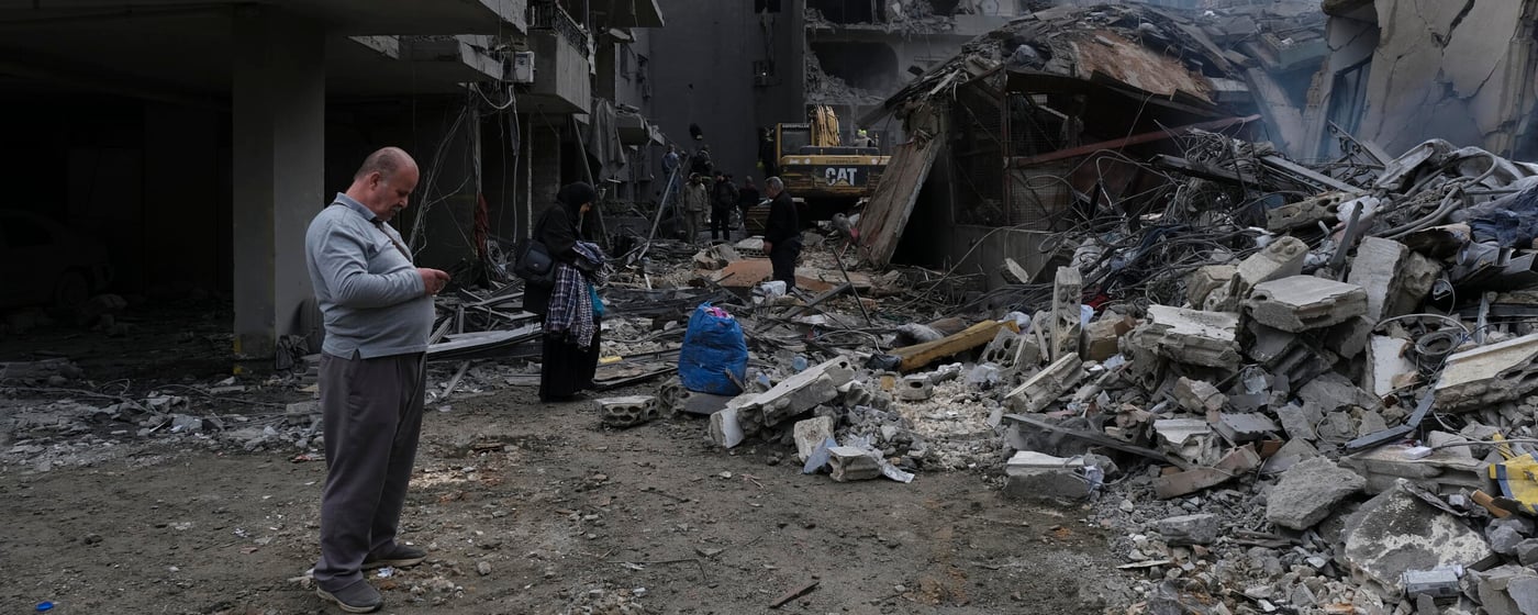 En gata ligger täckt av bråte efter att flera byggnader har rasat. Betong, metall och förstörda fasader visar spår av kraftigt våld. Människor rör sig försiktigt genom spillrorna, vissa bär på tillhörigheter medan andra ser ut att leta efter något användbart. En grävmaskin arbetar längre in i bildens mitt.
