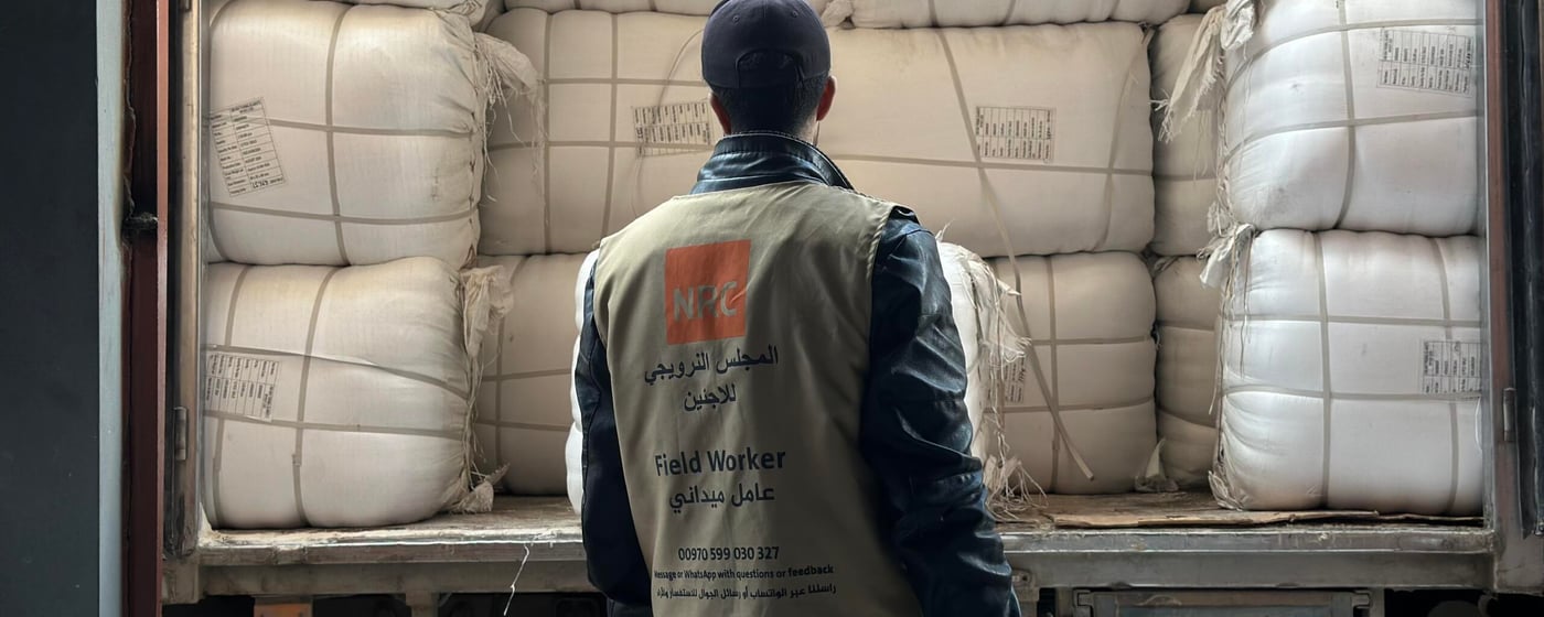 En man står bakom en lastbil lastad med stora vita säckar, staplade tätt intill varandra. Han bär en väst märkt med Norwegian Refugee Council och texten – Field Worker – och ser ut att inspektera eller förbereda lossning av humanitärt bistånd.
