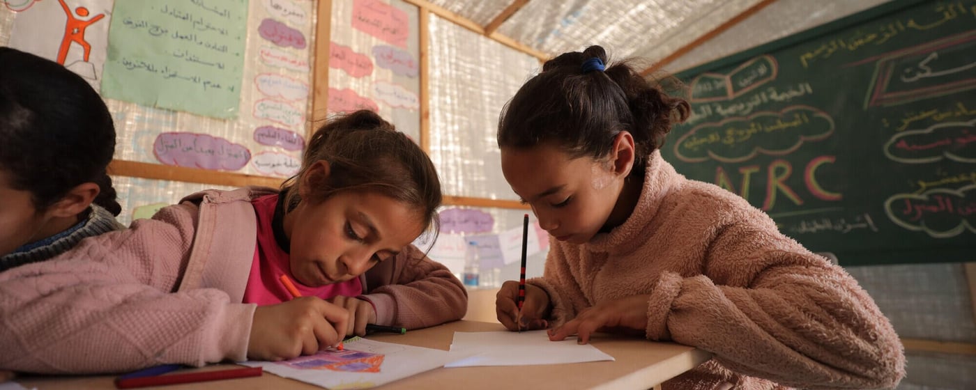 Två flickor sitter vid ett träbord i ett klassrum och ritar eller skriver på papper med färgpennor.