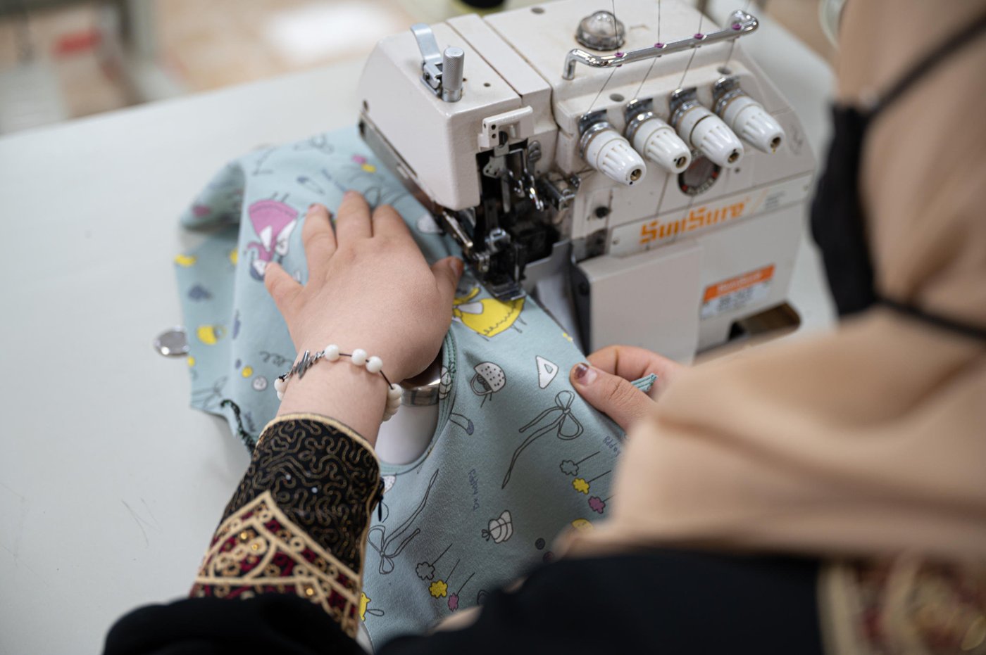 DO NOT USE (embargoed by head office)
Photo: Daniel Wheeler/NRC
Description: Ala (22) sewing some baby clothes as part of a UNICEF contract that the centre managed to secure.
Factual information
First name: Ala
Age: 22
Current location: Za’atari Refugee Camp, Jordan
From: Daraa, Syria. Fled to Jordan in 2012
Occupation: Seamstress at NRC’s youth programme.
NRC beneficiary: Yes, Ala received seamstress training as part of NRC’s youth training programme and now works as a seamstress as part of the programme.
Interview
Ala’s words
“I fled Syria in 2012 because of the heavy fighting. I left on foot at 10pm and arrived at the Jordanian border at 6am the next morning. We travelled at night because we were terrified of being attached on the way. There was around 100 of us and many people were injured.
When we arrived at the border and were met by the army (Jordanian Army) I felt like a drowning person that had been saved. I felt on edge the entire journey and now I had soldiers telling me ‘You are safe now’, it was all so overwhelming.
I live with my five sisters and five brothers. I am the eldest, so I feel responsible for them, and I looked after them during coronavirus.
I found out about NRC’s youth programme after doing a four-month sewing scholarship with a different organisation. I became very passionate about sewing so I wanted to continue my training and the programme at NRC seemed perfect. When I started on the programme, I realised that I still had so much to learn, and it was very intensive but rewarding.
The programme is delivered in three phases, with the first stages being training and the third phase being an employment opportunity.
I live on the other side of the camp and walk an hour each day to get to the centre. It’s a long walk in the desert heat but it’s so worth it.
An average day for me is waking up at 6am and after getting ready, I walk to the centre and we started work at 8:30am. At 12:30am we have a lunch break and then we work until 3pm. After I walk back home, I usually have a nap because the work is tiring but very rewarding.
When asked what the programme means to her, she replies with: “This opportunity that NRC has given me has provided me with financial independence and an opportunity for success in my life. My father is so proud of me and that is very important to me.”
When asked what she wants to do in the future, she says: “In the future, I want to open my own sewing centre for women which has integrity and quality – just like NRC.”
