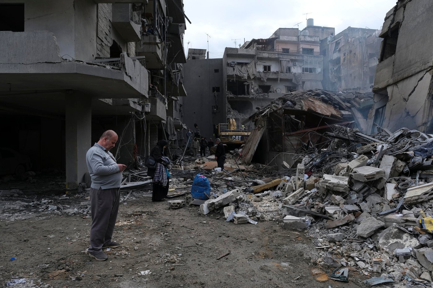 En gata ligger täckt av bråte efter att flera byggnader har rasat. Betong, metall och förstörda fasader visar spår av kraftigt våld. Människor rör sig försiktigt genom spillrorna, vissa bär på tillhörigheter medan andra ser ut att leta efter något användbart. En grävmaskin arbetar längre in i bildens mitt.