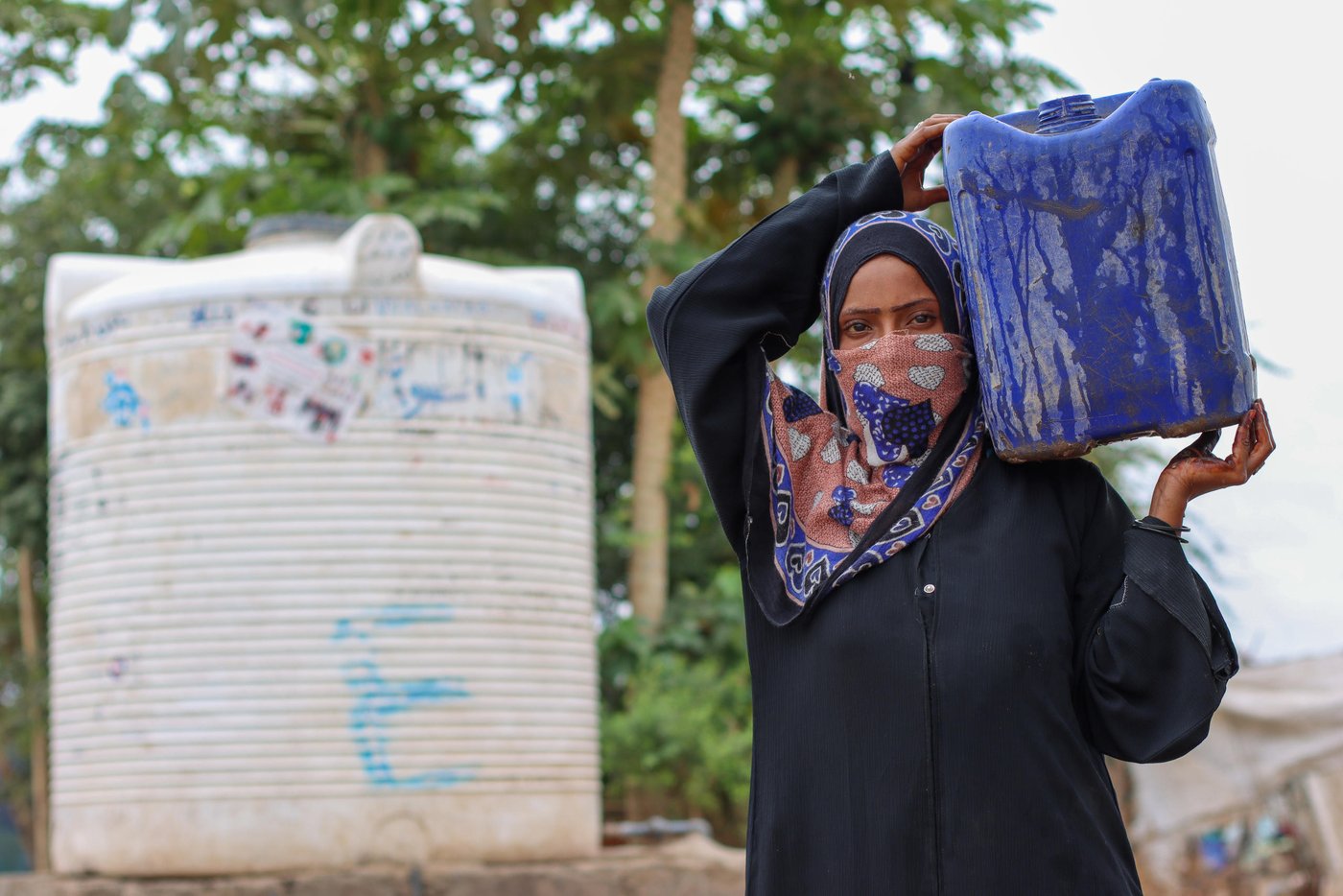 A woman carrying a blue water can on her left shoulder. In the background we see the large, white tank where she collected the water. 