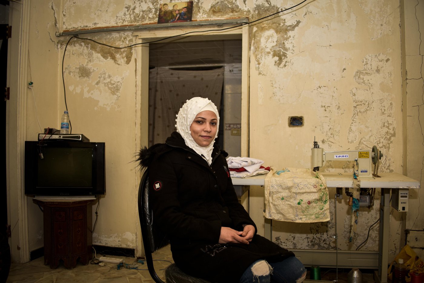 A woman sitting in her working space where she keeps her sewing machine and fabrics.