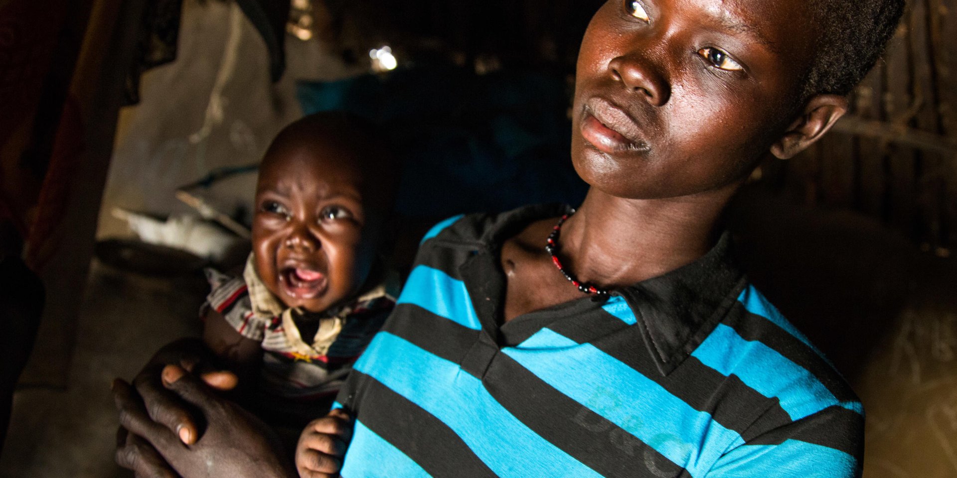 8 March 2017. Ngop: Marsa Nyalaih holds her baby in their shelter in Ngop, Unity state, South Sudan. The house hosts ten children and the family has difficulties to feed them all.

Marsa is a mother of three children, but in the house there are nearly 20 family members, half of them children. “We are really struggling to feed all of them,” Marsa explains. That’s why they even started eating those maize seeds that humanitarian organizations distributed last year for cultivation. “Last year, our land didn’t produce much and we don’t want to risk and sow again for another bad season,” Marsa explains while she is boiling a big pot of seeds. “If we plant these seeds, we lose the opportunity to have something to eat these difficult days,” she adds.

Her mother Nyakur Deng regrets the tough life in Ngop. “I feel sad seeing my daughter having so many troubles to feed my grandchildren,” Nyakur says while holding one of them. 

Wild fruits and tree leaves are other options to sustain the family, but Marsa explains that the nearest places to collect them are three hours away from the house. “We need to walk long distances to just have some vegetables that are not even enough to satisfy my children,” Marsa concludes. 

The Norwegian Refugee Council (NRC) distributed food (maize, lentils, oil and corn soya blend) for more than 7,100 people in Ngop. While Leer and Mayendit, both also in Unity state, have been recently declared in famine, Ngop is also in high risk. The food distribution is part of a Rapid Response Mission coordinated and funded by the World Food Programme (WFP) to mitigate this risk. 
Photo by Albert Gonzalez Farran - NRC