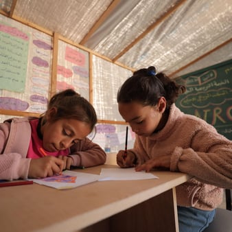 Två flickor sitter vid ett träbord i ett klassrum och ritar eller skriver på papper med färgpennor.