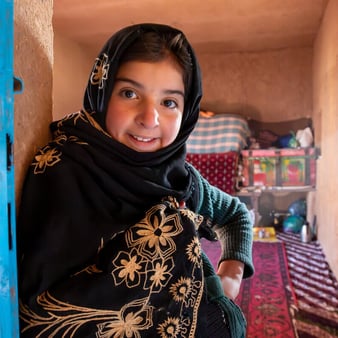 En ung flicka står i dörröppningen till sitt hem och ler mot kameran. Hon bär en svart sjal med broderade blommor i guld och en grön tröja.