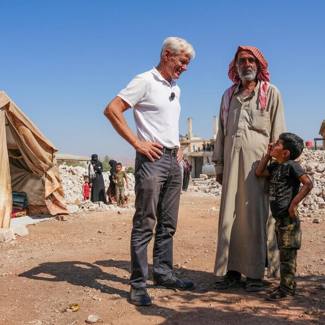 En man från Norwegian Refugee Council står och samtalar med en äldre man och ett barn i ett område med tält och förstörda byggnader. Bakom dem syns fler människor och rester av hus i det torra landskapet under en klar himmel.