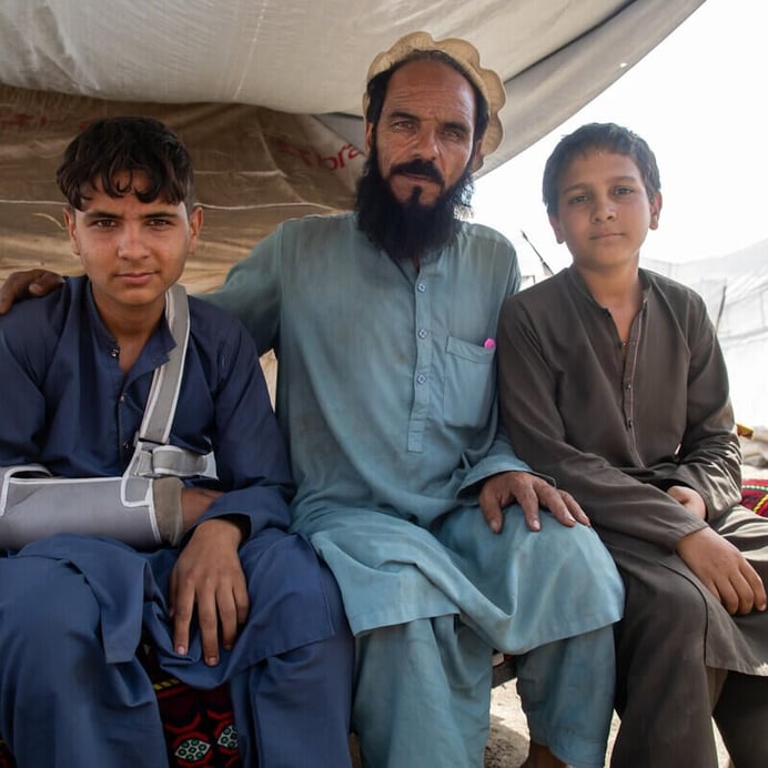 En man sitter i ett tält tillsammans med två pojkar. En av pojkarna har en armskena.  De befinner sig i ett tillfälligt boende efter jordbävningen.