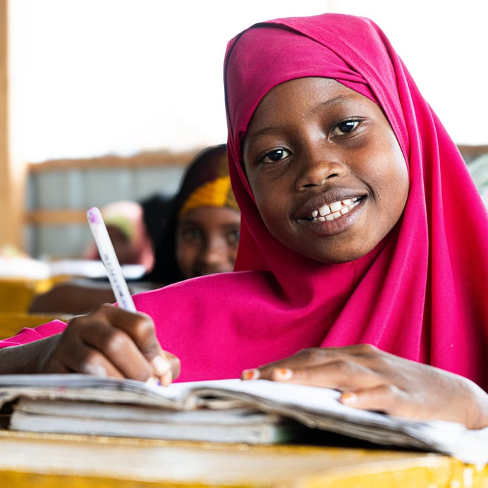 En flicka sitter vid en skolbänk i ett klassrum i Towfiq-lägret i Jowhar. Hon ler mot kameran medan hon skriver i sitt anteckningsblock. Ljuset faller in från sidan och andra elever syns i bakgrunden.