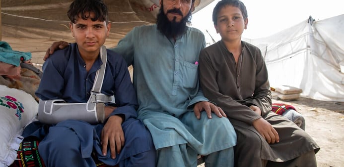 En man sitter i ett tält tillsammans med två pojkar. En av pojkarna har en armskena.  De befinner sig i ett tillfälligt boende efter jordbävningen.