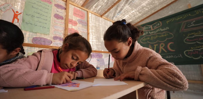 Två flickor sitter vid ett träbord i ett klassrum och ritar eller skriver på papper med färgpennor.