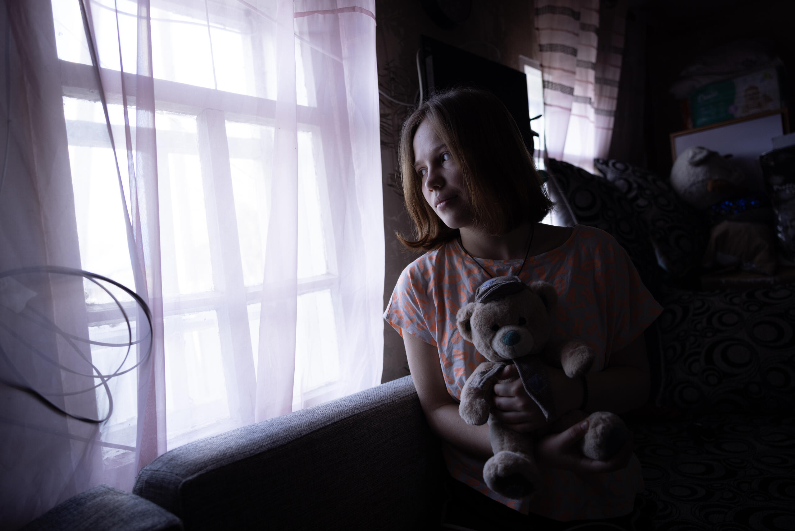 En flicka st&aring;r vid ett f&ouml;nster i sitt hem i Ukraina och h&aring;ller en nallebj&ouml;rn. Kriget forts&auml;tter att p&aring;verka barns vardag och trygghet.