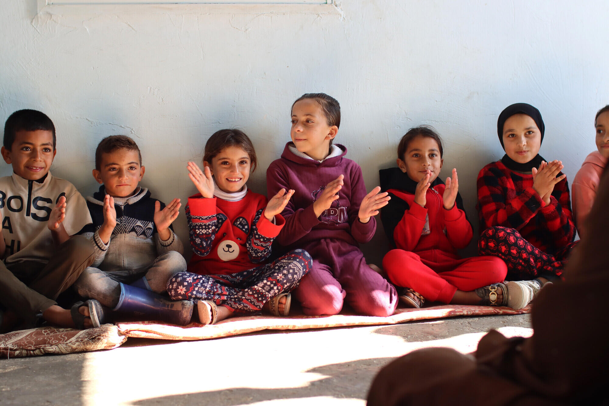 En kvinna sitter p&aring; marken utomhus tillsammans med flera barn och visar dem hur de ska arbeta med papper och pennor.