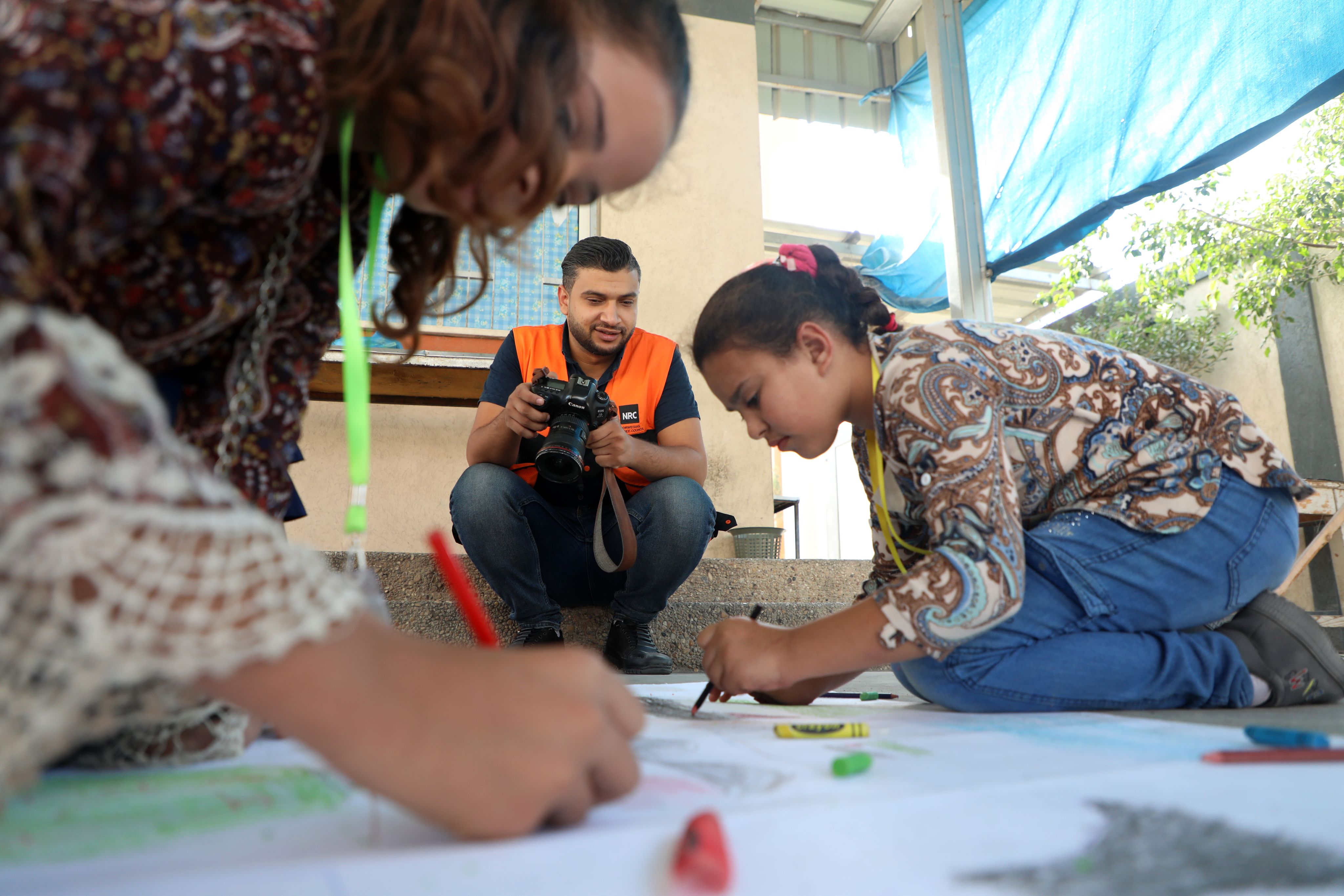 Yousef, en mann med mørkt hår og oransje vest, fotograferer to elever som tegner på ark på bakken.