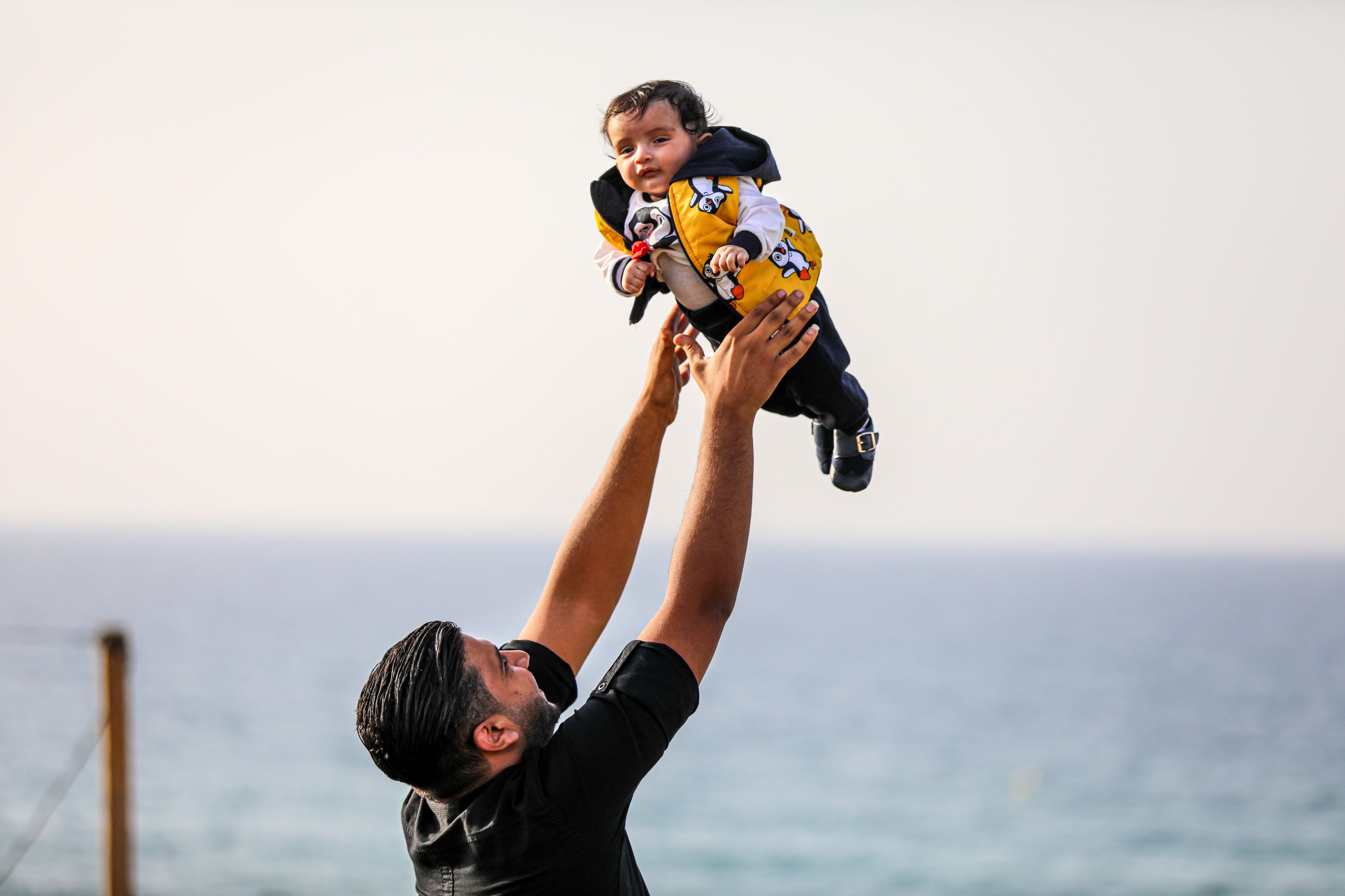 Yousef leker med sønnen sin ved å kaste han opp i luften. Den lille kutten hat på seg en gul jakke med pingvinmønster.