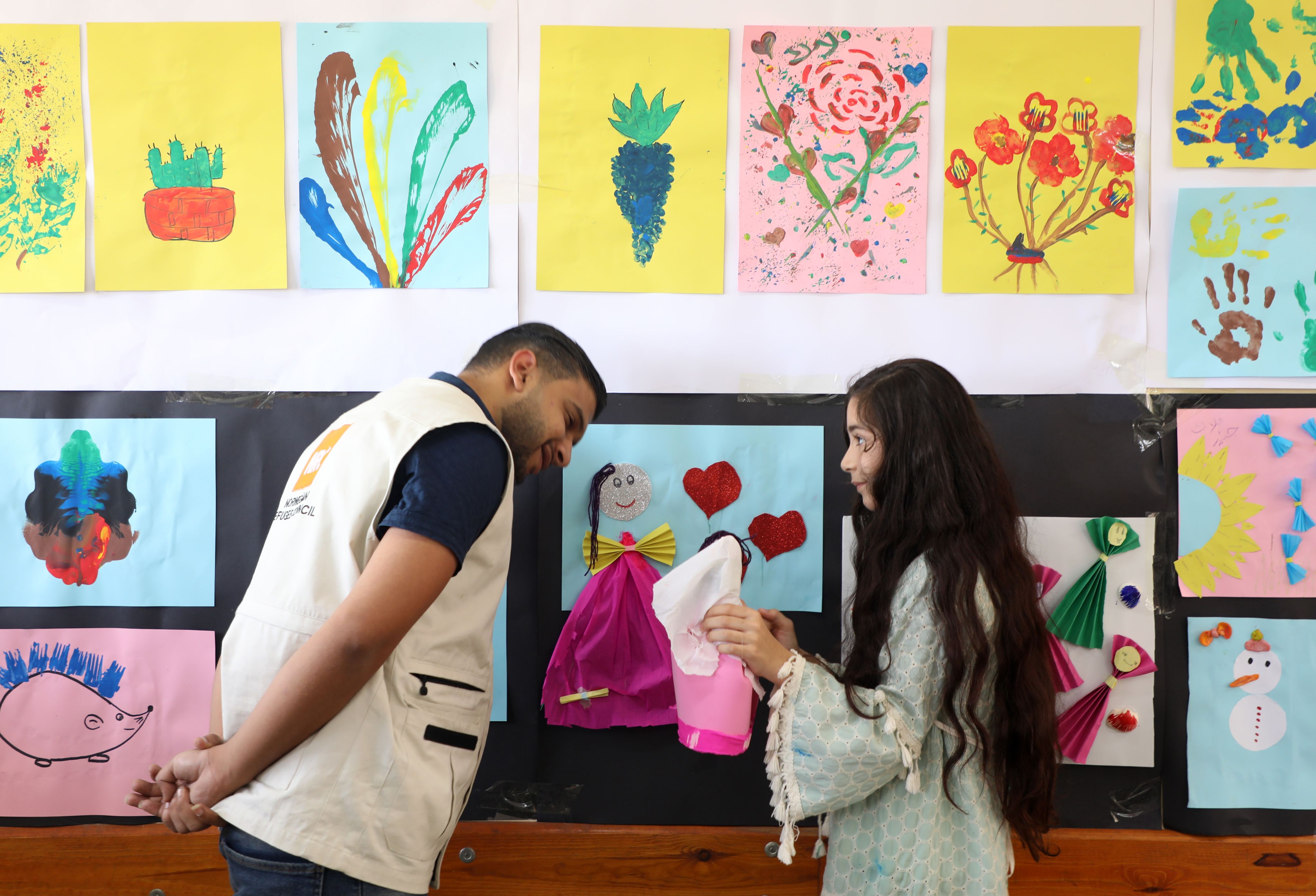 Yousef i prat med en elev. Hun viser ham noe hun har laget. Veggen bak dem er full av fargerike tegninger.