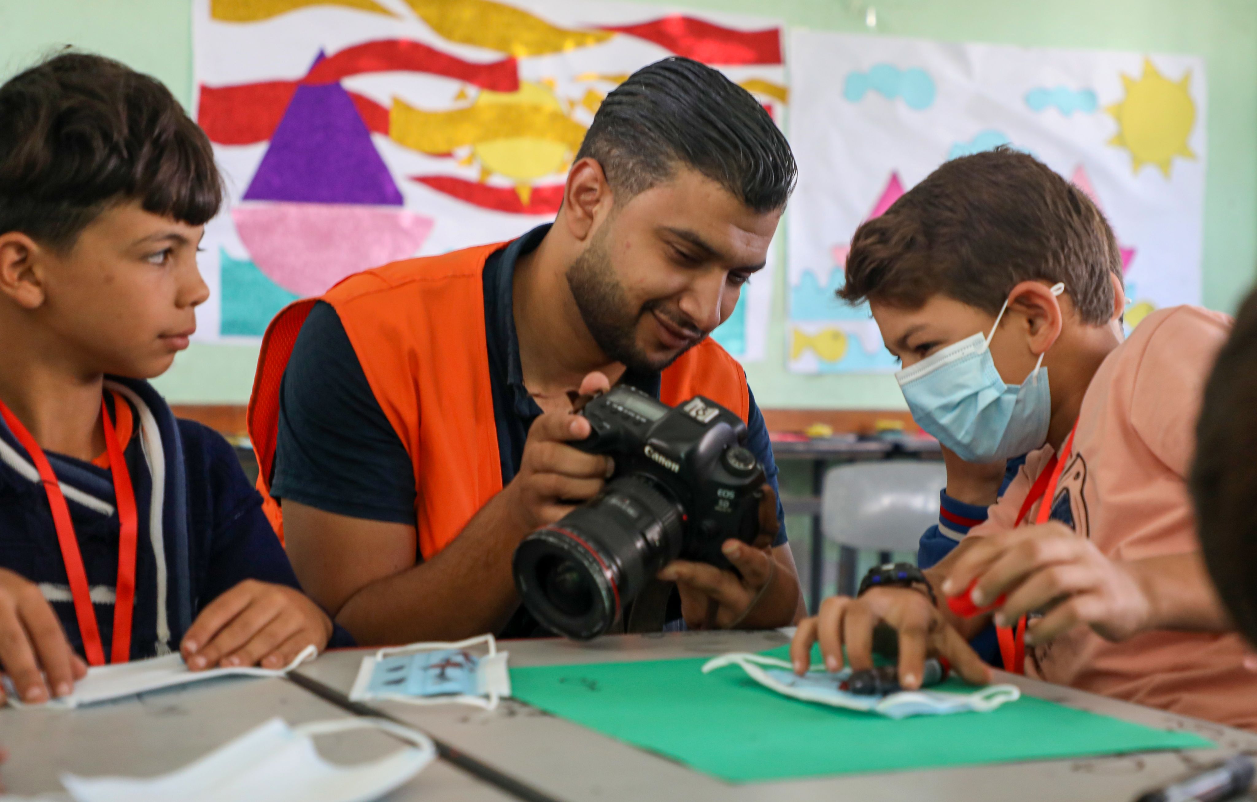Yousef, en mann med mørk hår og oransje vest, viser to barn et bilde på kameraet sitt.