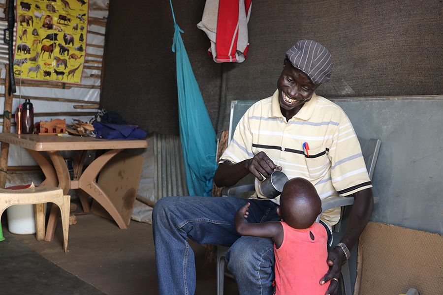 A father, with a bog smile on his face, is giving his daughter water. He is wearing a six pence and a striped t-shirt, his daughter a bright pink shirt.