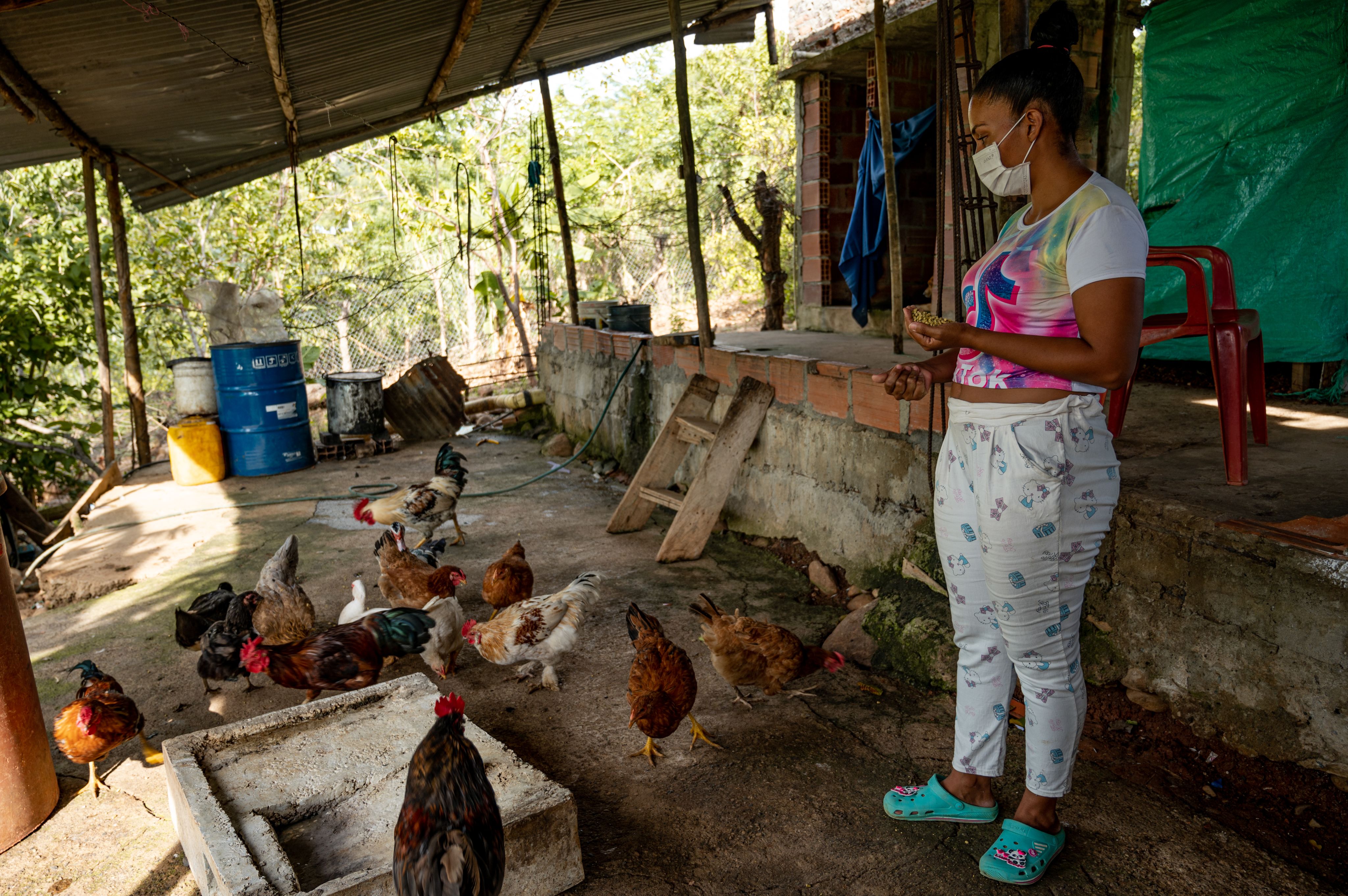 A woman in white trousers and a colourful top is feeding chickens. They're standing on a concrete floor.