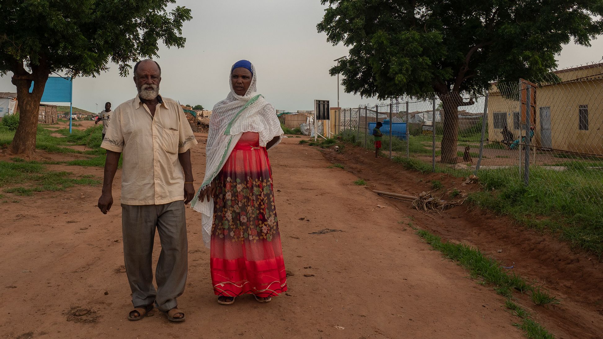 An elderly man and woman walking next to each other on a dirt road. Two large trees and some low buildings line the sides of the road.
