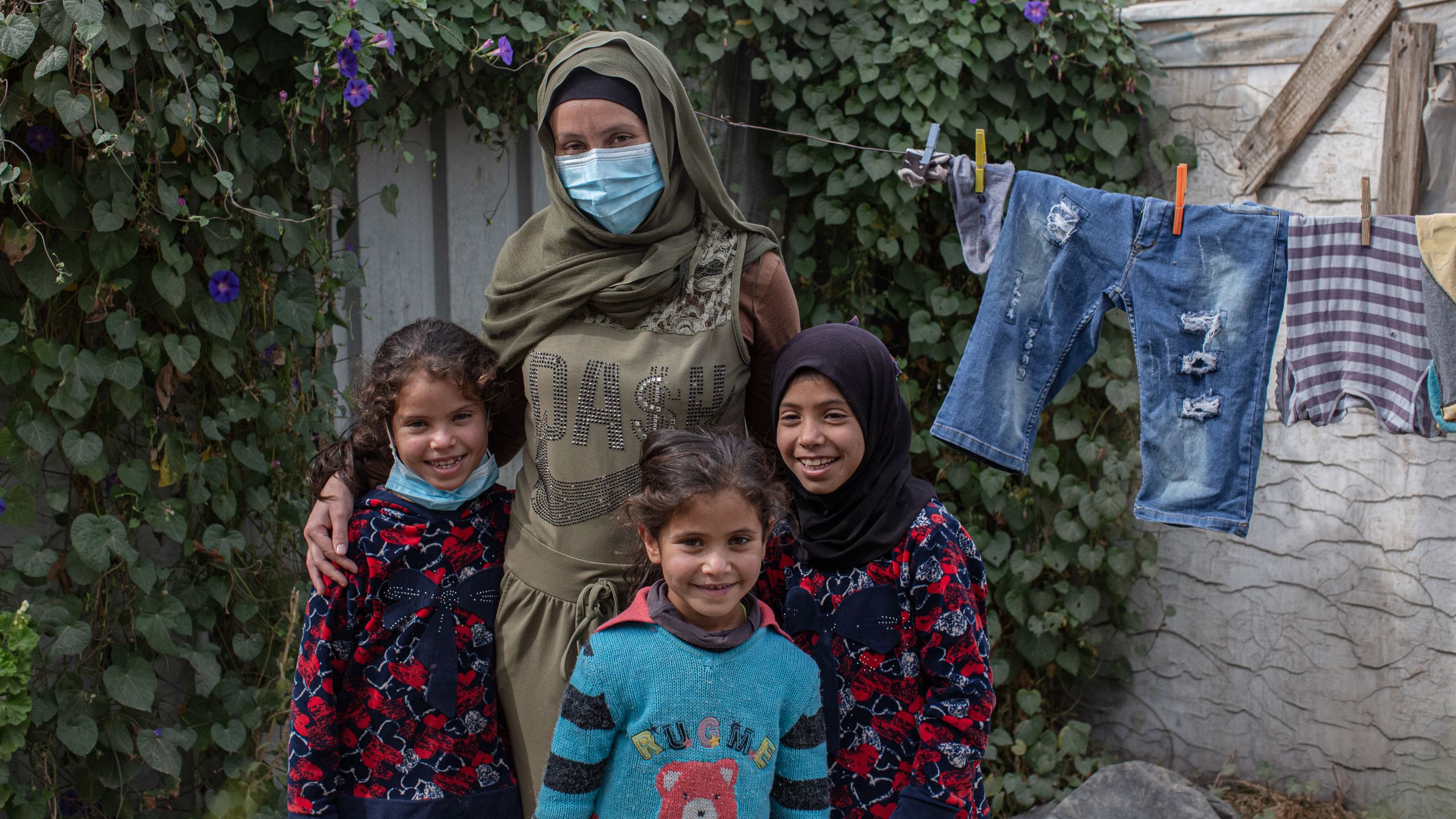 A woman and her three daughters stand outside their tent. The walls are covered in climbing plants. Laundry hangs on a clothesline behind them.