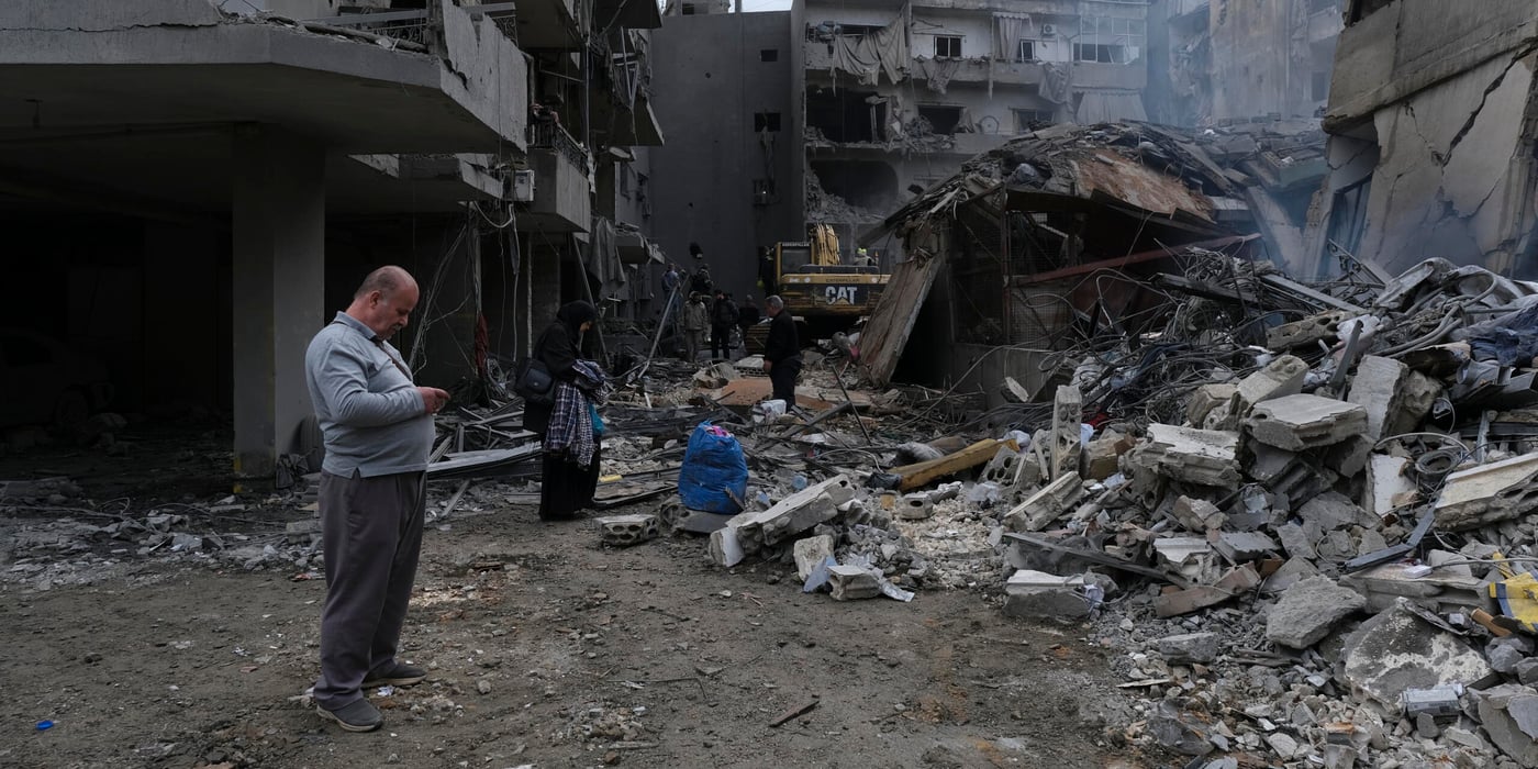En gata ligger täckt av bråte efter att flera byggnader har rasat. Betong, metall och förstörda fasader visar spår av kraftigt våld. Människor rör sig försiktigt genom spillrorna, vissa bär på tillhörigheter medan andra ser ut att leta efter något användbart. En grävmaskin arbetar längre in i bildens mitt.