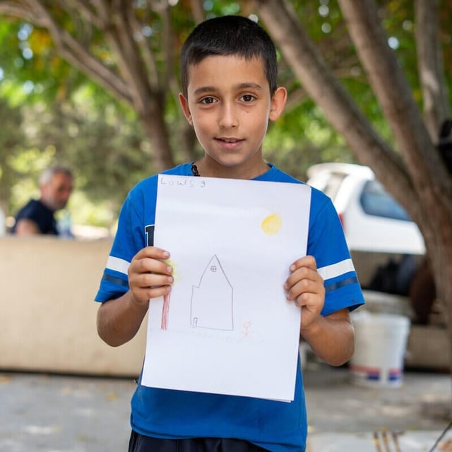 En pojke står utomhus och håller upp en teckning av ett hus med sol och en person. I bakgrunden syns träd, några vuxna och parkerade fordon.